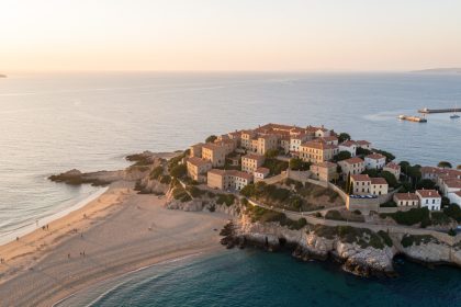 Vue aérienne réaliste de Fos-sur-Mer au lever du soleil, avec une plage de sable, un vieux village sur une légère hauteur et la mer Méditerranée baignée de lumière provençale.