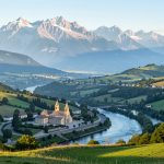 Vue panoramique lumineuse de Lourdes depuis les hauteurs, avec le sanctuaire Notre-Dame de Lourdes, le Gave de Pau et les montagnes pyrénéennes en arrière-plan.
