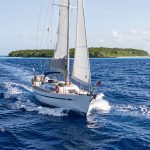 Voilier hauturier naviguant sur une mer bleu profond du Pacifique Est vers un atoll tropical isolé, dans une ambiance réaliste de préparation d’expédition.