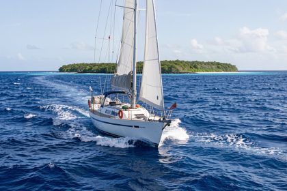Voilier hauturier naviguant sur une mer bleu profond du Pacifique Est vers un atoll tropical isolé, dans une ambiance réaliste de préparation d’expédition.