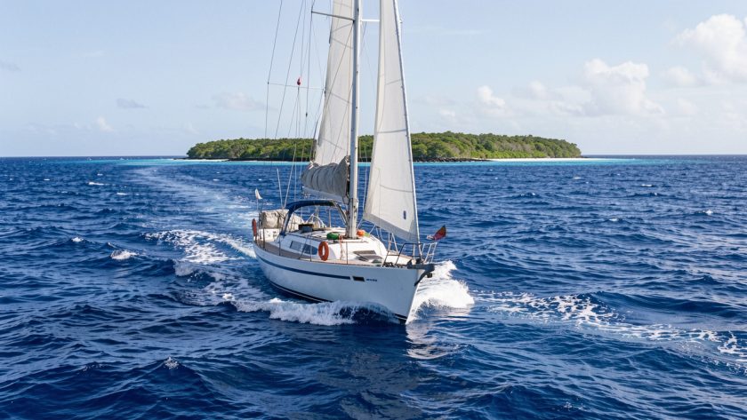 Voilier hauturier naviguant sur une mer bleu profond du Pacifique Est vers un atoll tropical isolé, dans une ambiance réaliste de préparation d’expédition.