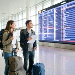 Trois voyageurs avec des bagages souples et des documents de croisière à voile devant un panneau lumineux dans un aéroport moderne.